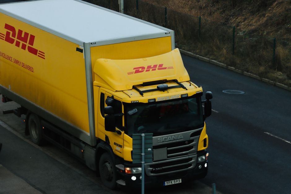 Caminhão de entrega amarelo da DHL dirigindo por uma estrada da cidade, mostrando a logística em ação.