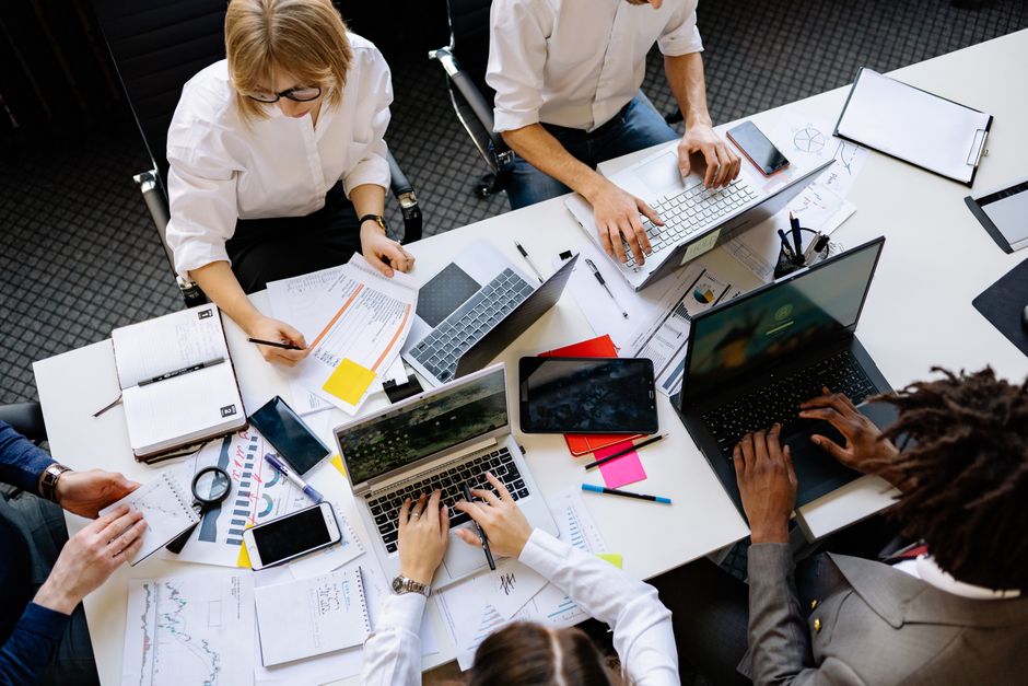 Cena vibrante de trabalho em equipe no escritório com laptops, documentos e profissionais diversos em uma reunião.