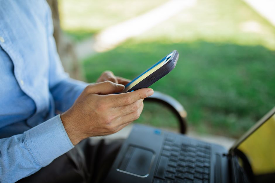 Uma pessoa usa um smartphone enquanto está sentada ao ar livre com um laptop, demostrando o estilo de vida do trabalho remoto.