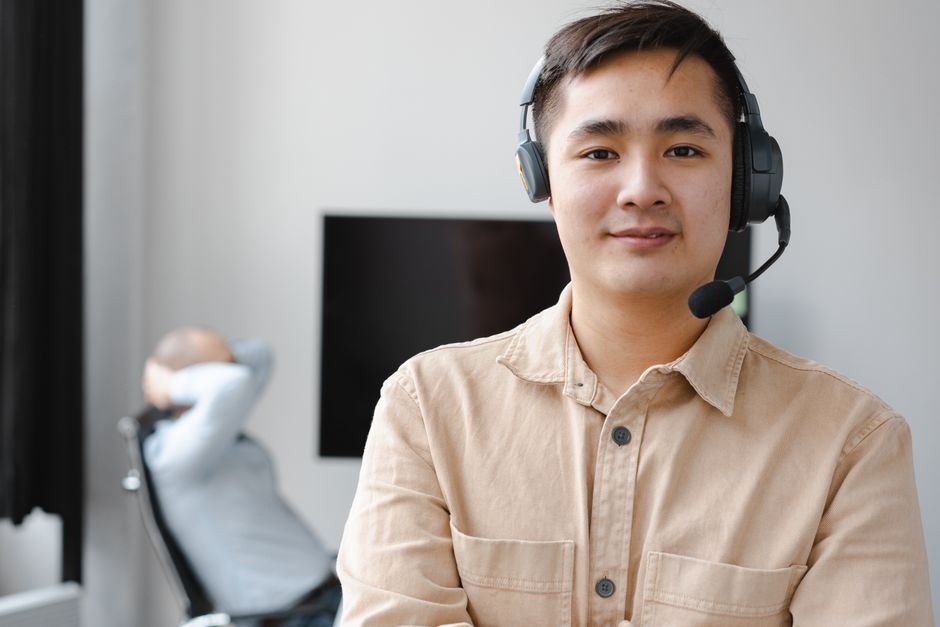 Homem asiático vestindo uma camisa marrom e fones de ouvido sorri em um ambiente de escritório moderno.