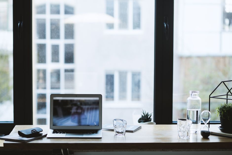 Configuração elegante de mesa de escritório com um laptop, copo de água e vista para a janela aberta.