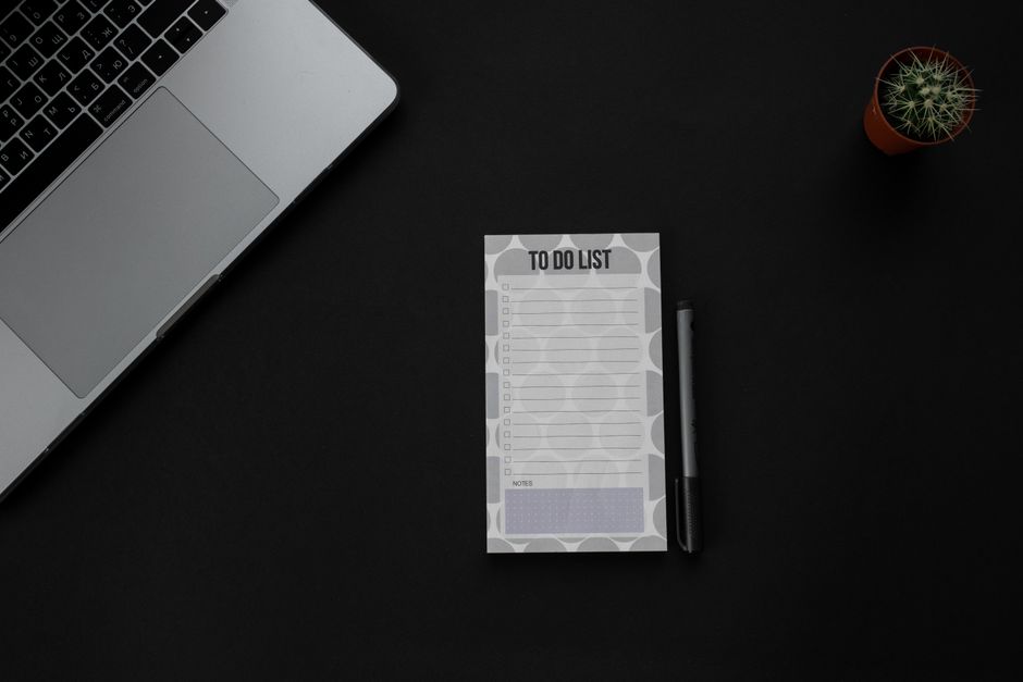 Um setup de mesa minimalista com um laptop, lista de tarefas e caneta sobre um fundo escuro. Ideal para temas de produtividade.